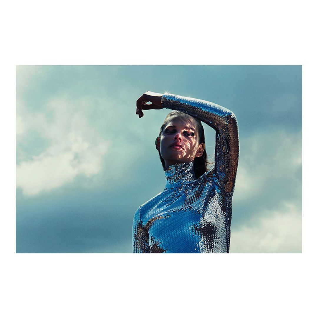 Young woman wearing a metallic silver dress poses outdoors under a cloudy sky, creating a futuristic and high-fashion aesthetic.