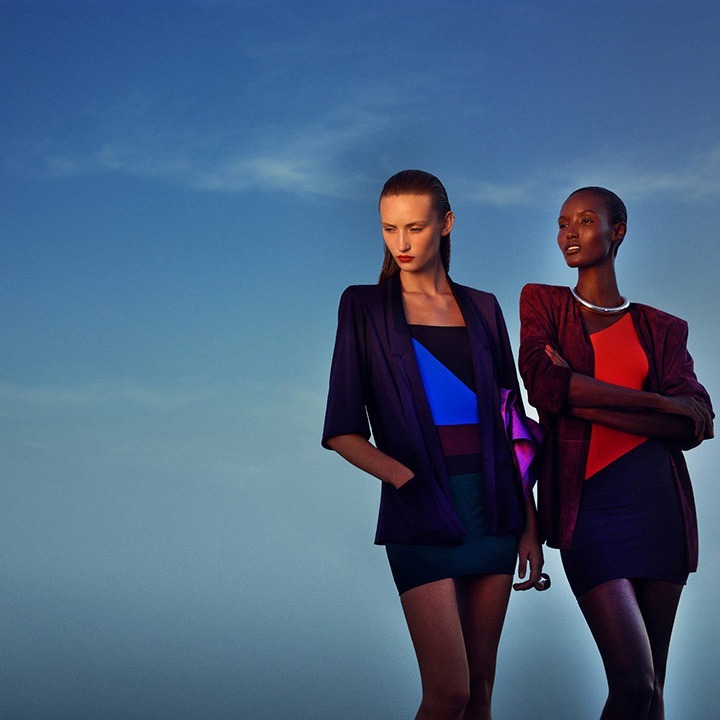 Two fashion models in modern dresses and blazers pose against a blue sky, creating a stylish and contemporary outdoor fashion shot.