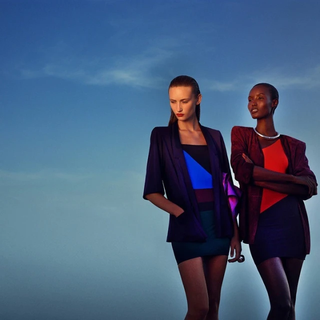 Two fashion models in modern dresses and blazers pose against a blue sky, creating a stylish and contemporary outdoor fashion shot.