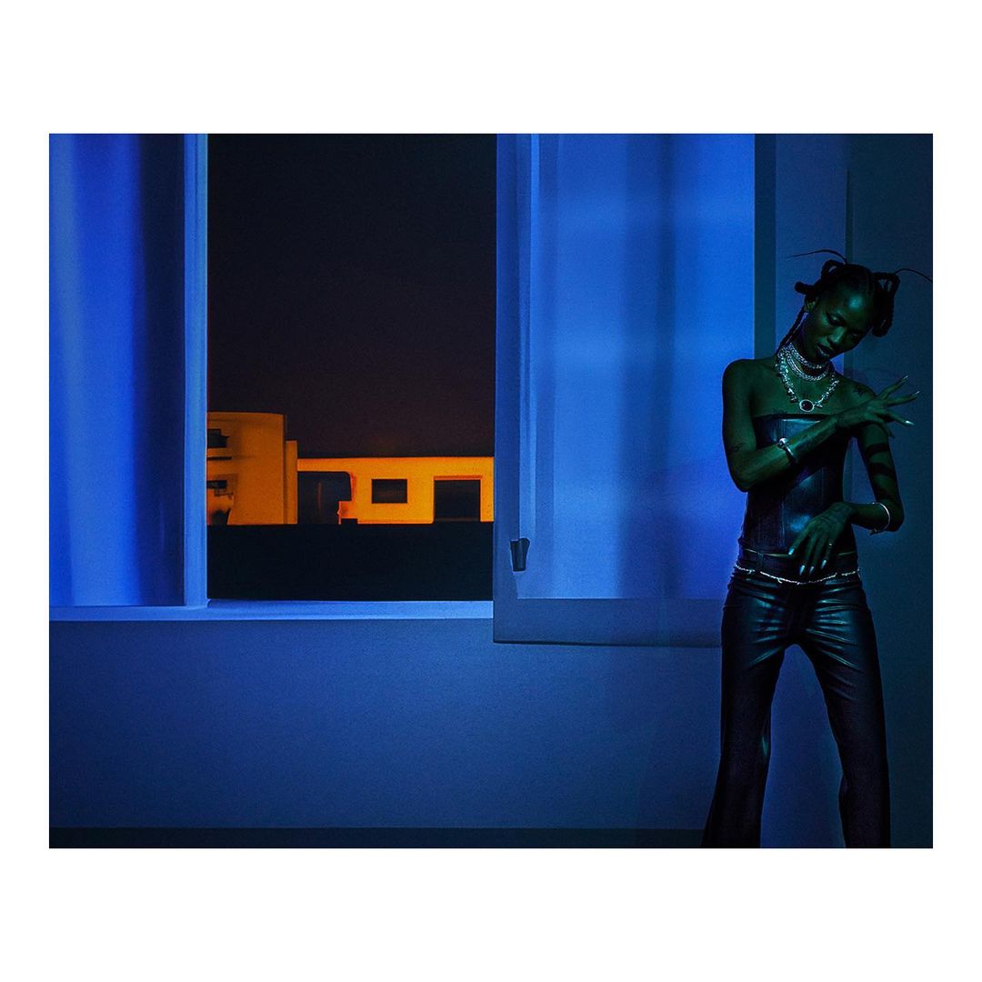 Elegant woman in black leather outfit strikes a pose indoors by a blue-lit window, offering a contemporary fashion statement.
