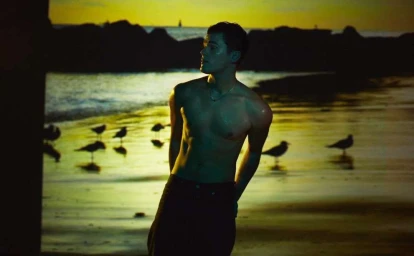 Shirtless young man standing on a sandy beach at sunset with seabirds foraging in the background.