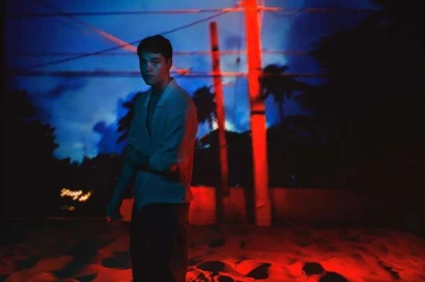 Young man in an open-collared shirt standing outdoors at night under dramatic red and blue lighting by Luis Sanchis.