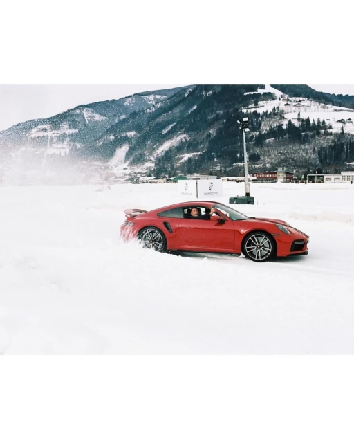 Red Porsche 911 Turbo S drives through snowy terrain, surrounded by snow drifts with snow-covered mountains in the background on a cloudy day.