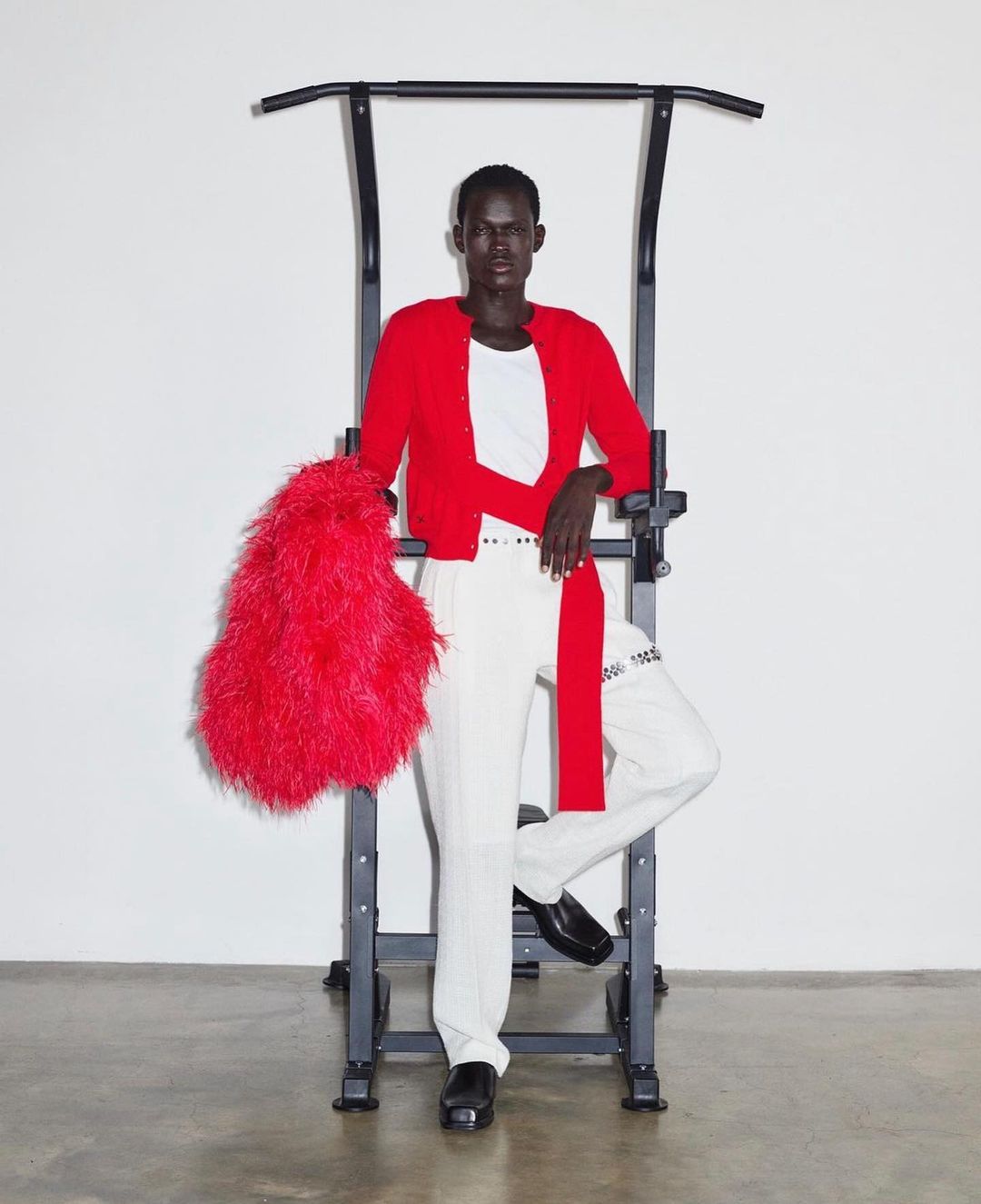 Dark-skinned adult male in a red cardigan, white pants, and black shoes poses indoors with a black pull-up bar and a red feathered bag.