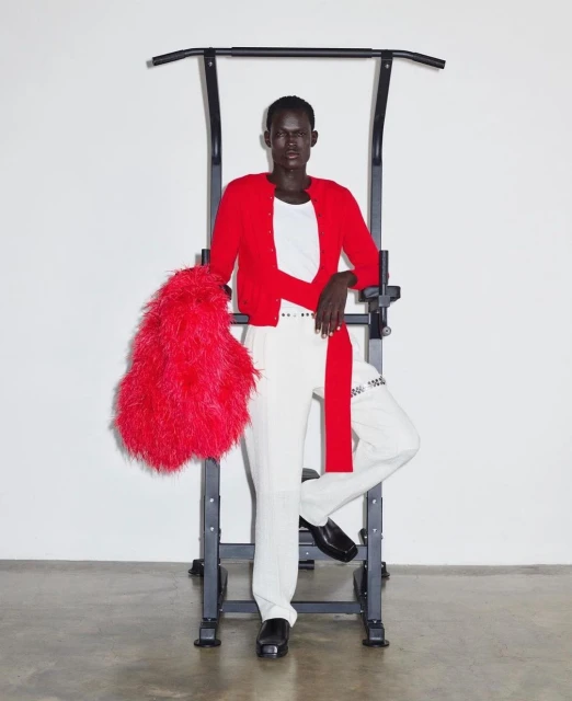 Dark-skinned adult male in a red cardigan, white pants, and black shoes poses indoors with a black pull-up bar and a red feathered bag.