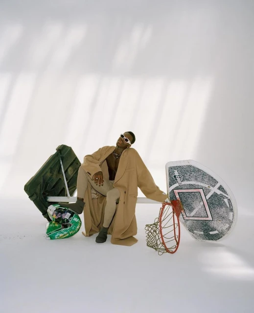 Stylish person poses in studio, wearing sunglasses and a long coat, seated next to a basketball hoop and helmet in a well-lit shot.