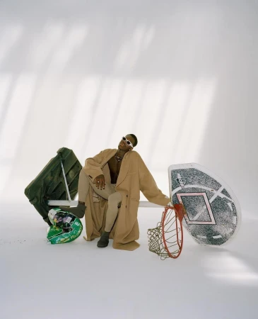 Stylish person poses in studio, wearing sunglasses and a long coat, seated next to a basketball hoop and helmet in a well-lit shot.
