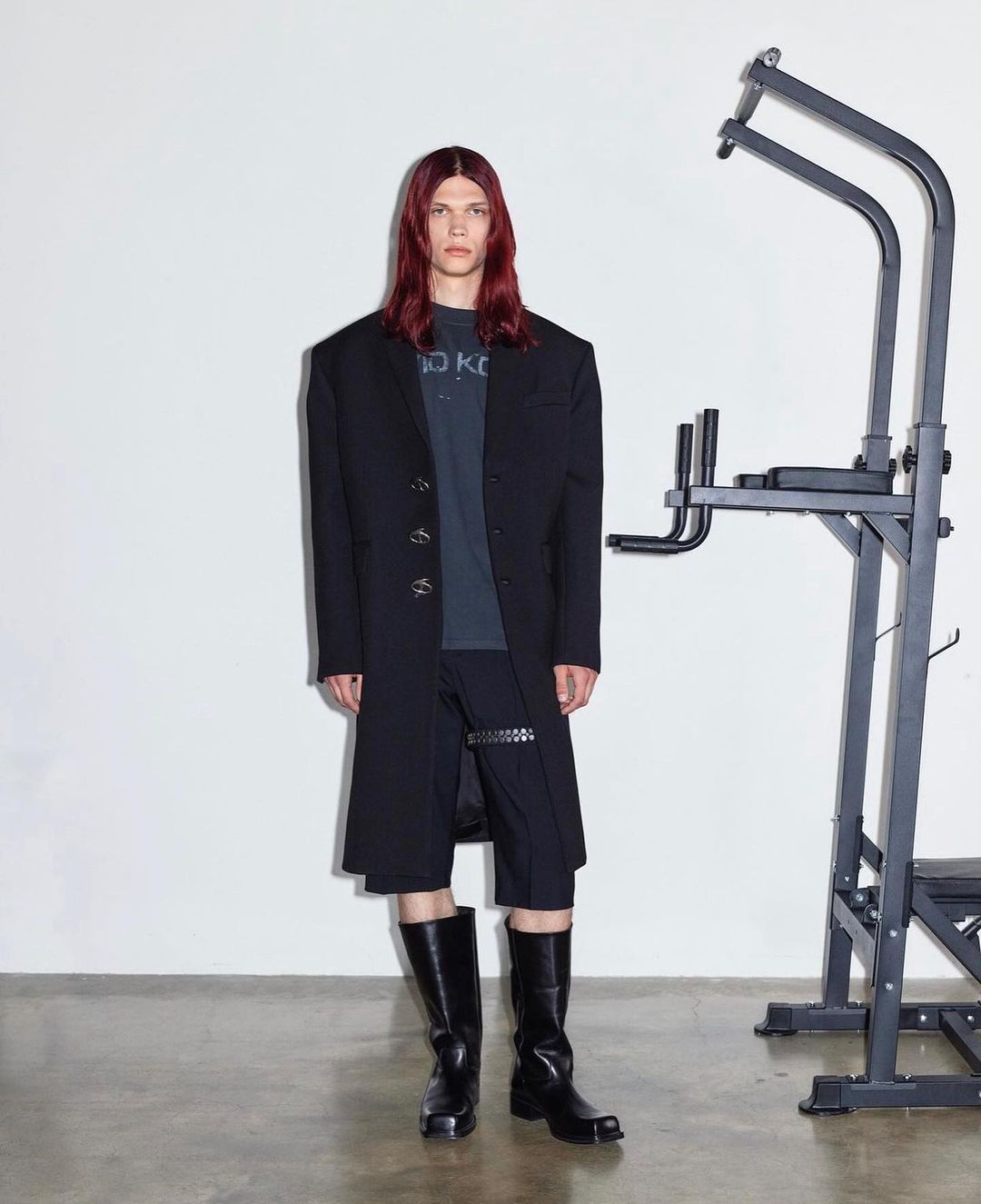 A model poses indoors wearing a black overcoat, shorts, and boots, with a pull-up bar in the background against a white wall.