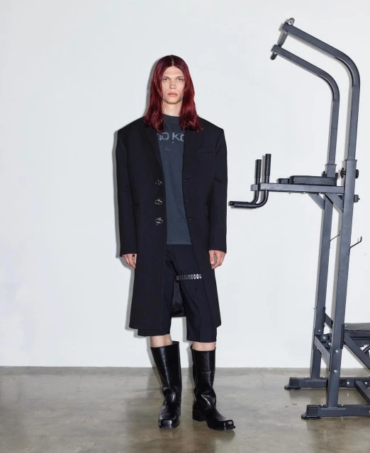 A model poses indoors wearing a black overcoat, shorts, and boots, with a pull-up bar in the background against a white wall.