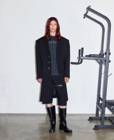 A model poses indoors wearing a black overcoat, shorts, and boots, with a pull-up bar in the background against a white wall.