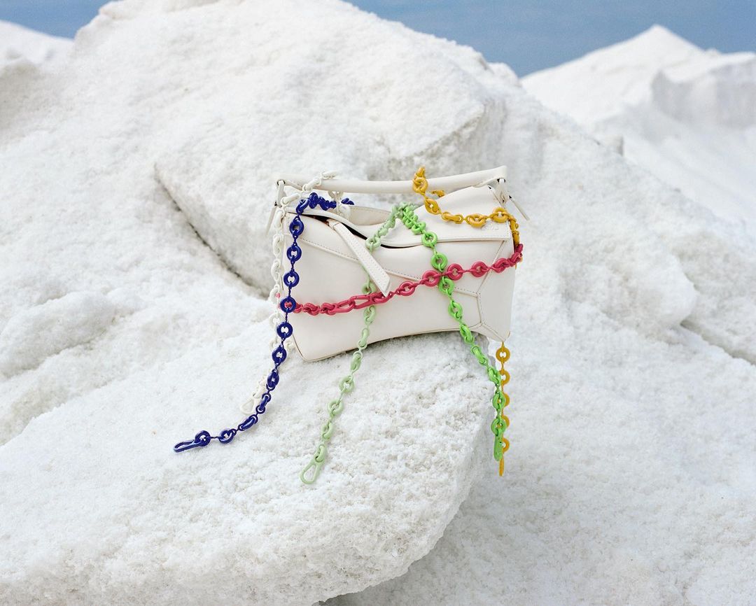 Fashion product shot of a white leather purse with colorful chain straps, posed on textured white rock in a bright, outdoor setting.