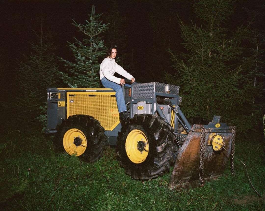 An adult posing on a yellow heavy equipment vehicle in an outdoor, nighttime setting surrounded by pine trees.