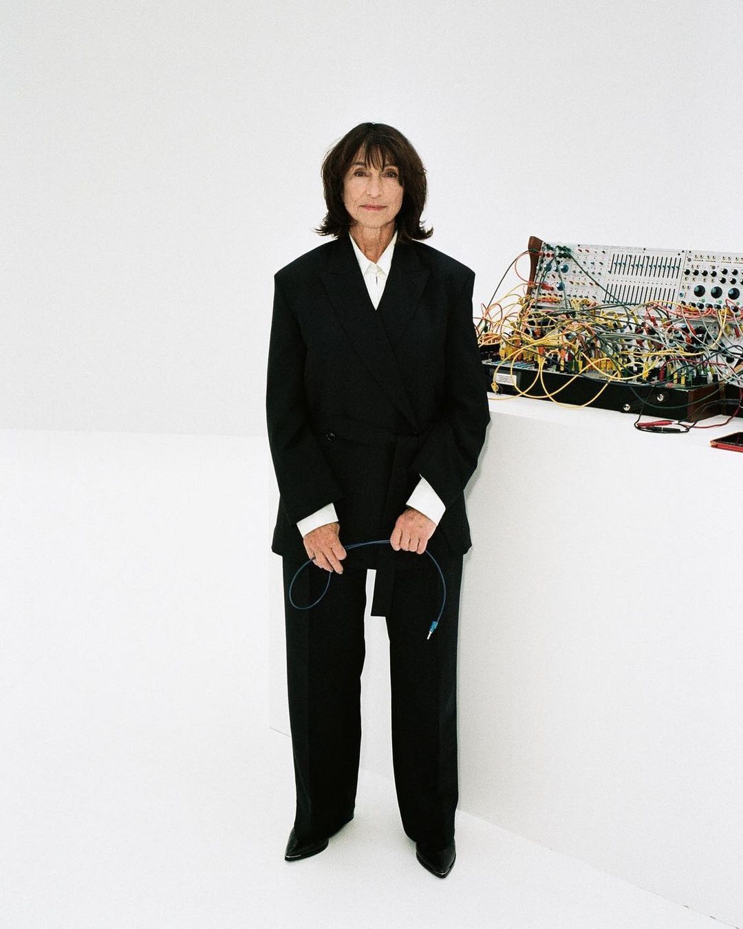 Mature woman in a black suit holding a cable, standing in front of a modular synthesizer on a white backdrop in a portrait orientation.