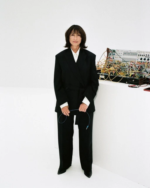 Mature woman in a black suit holding a cable, standing in front of a modular synthesizer on a white backdrop in a portrait orientation.