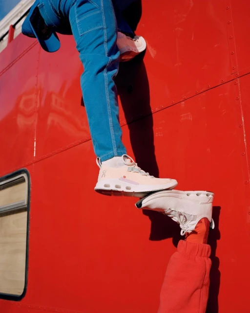 Two people display white ON brand sneakers against a red surface. One person wears blue pants; the other, red sweatpants.