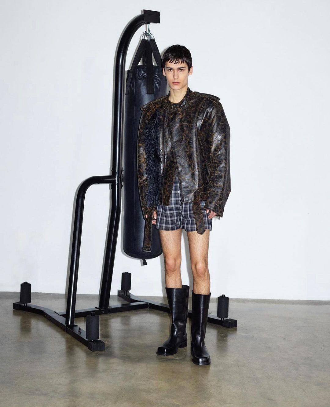 Adult male poses indoors near a black punching bag, wearing a distressed leather jacket, plaid shorts, and black boots on a concrete floor.