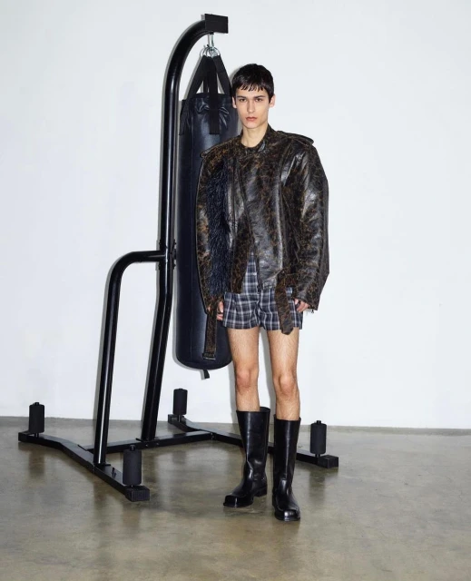 Adult male poses indoors near a black punching bag, wearing a distressed leather jacket, plaid shorts, and black boots on a concrete floor.
