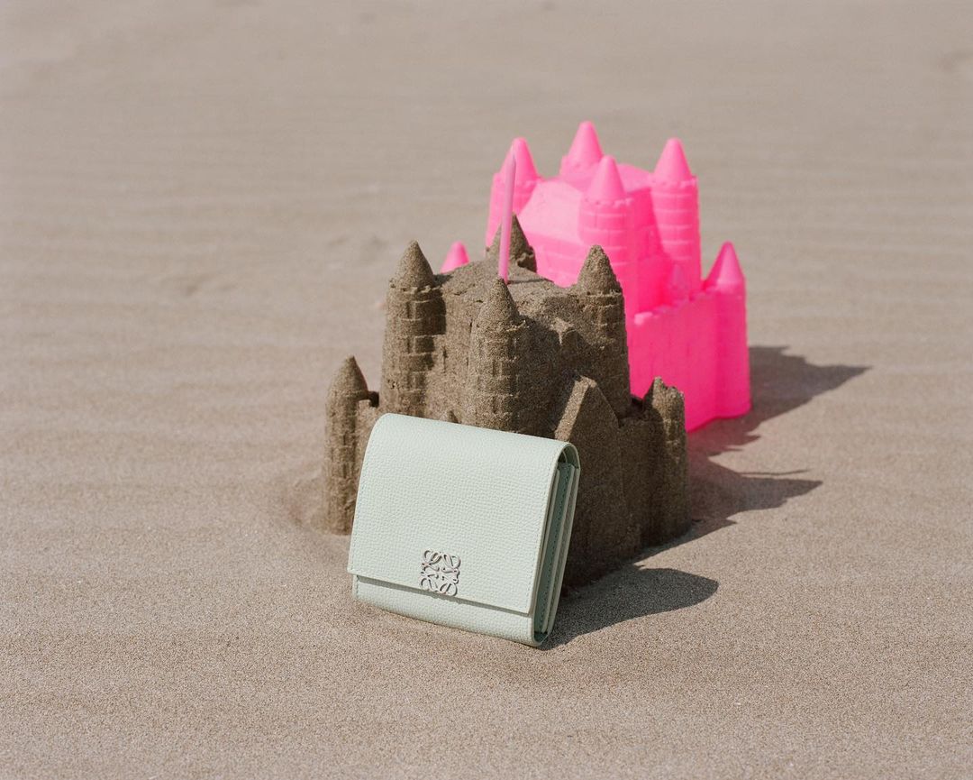 Light green wallet sits on a sandy beach with pink and brown sandcastles in the background, illuminated by natural light.