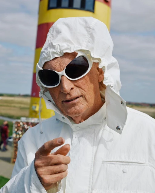 An older adult in a white hooded jacket and sunglasses gestures toward the camera near a red-and-yellow striped tower on a sunny day.