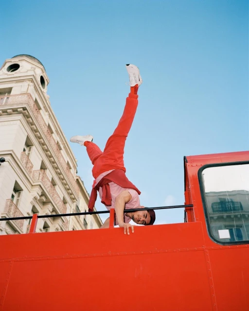 Young adult doing a handstand on the top of a red double-decker bus with a building and blue sky in the background. 