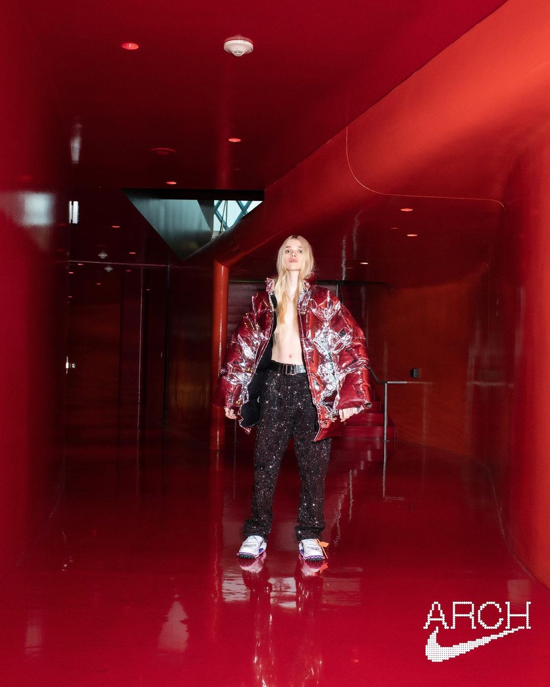 Fashion-forward adult wearing a bold jacket and patterned pants, posing in a sleek red hallway with a futuristic design.
