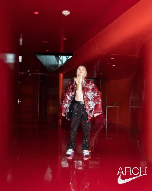 Fashion-forward adult wearing a bold jacket and patterned pants, posing in a sleek red hallway with a futuristic design.