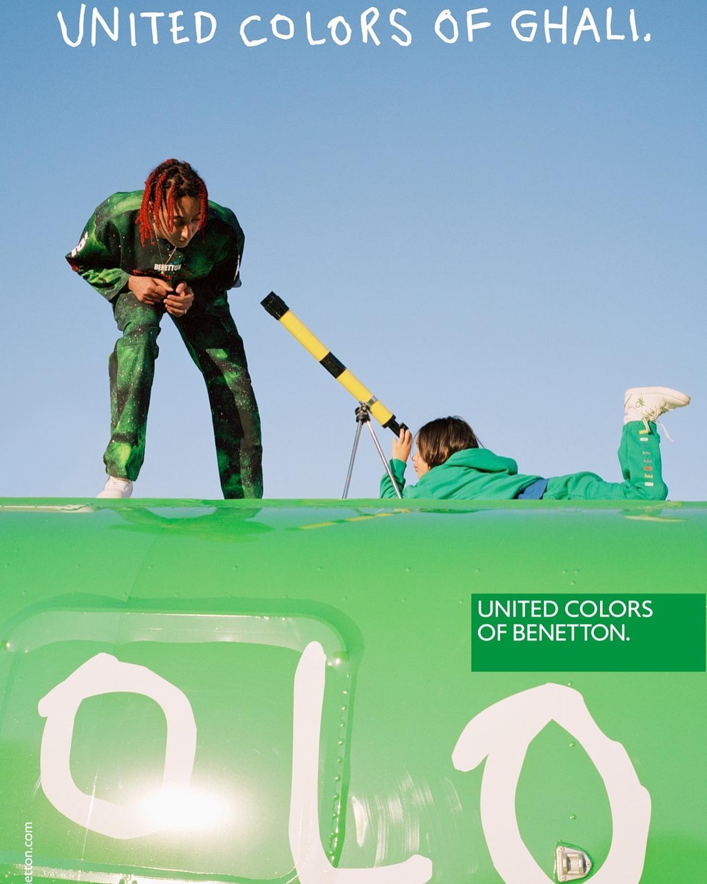 Two people on a bright green surface against a clear blue sky: an adult standing, and a child looking through a yellow telescope.