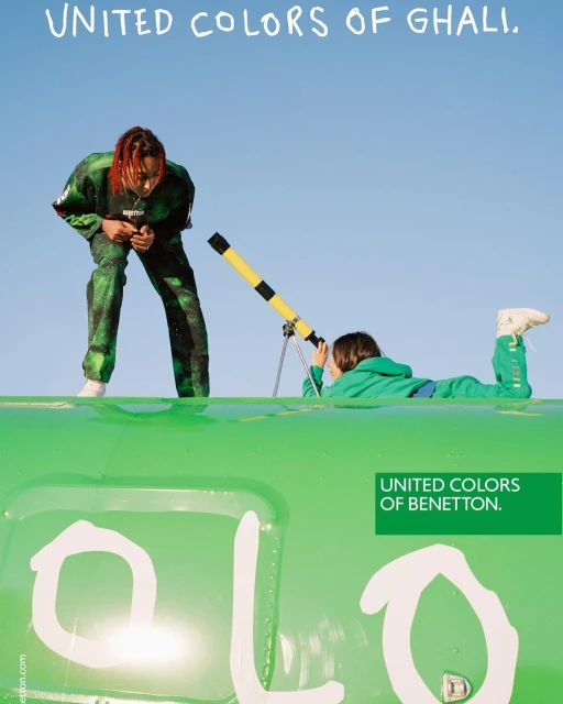 Two people on a bright green surface against a clear blue sky: an adult standing, and a child looking through a yellow telescope.