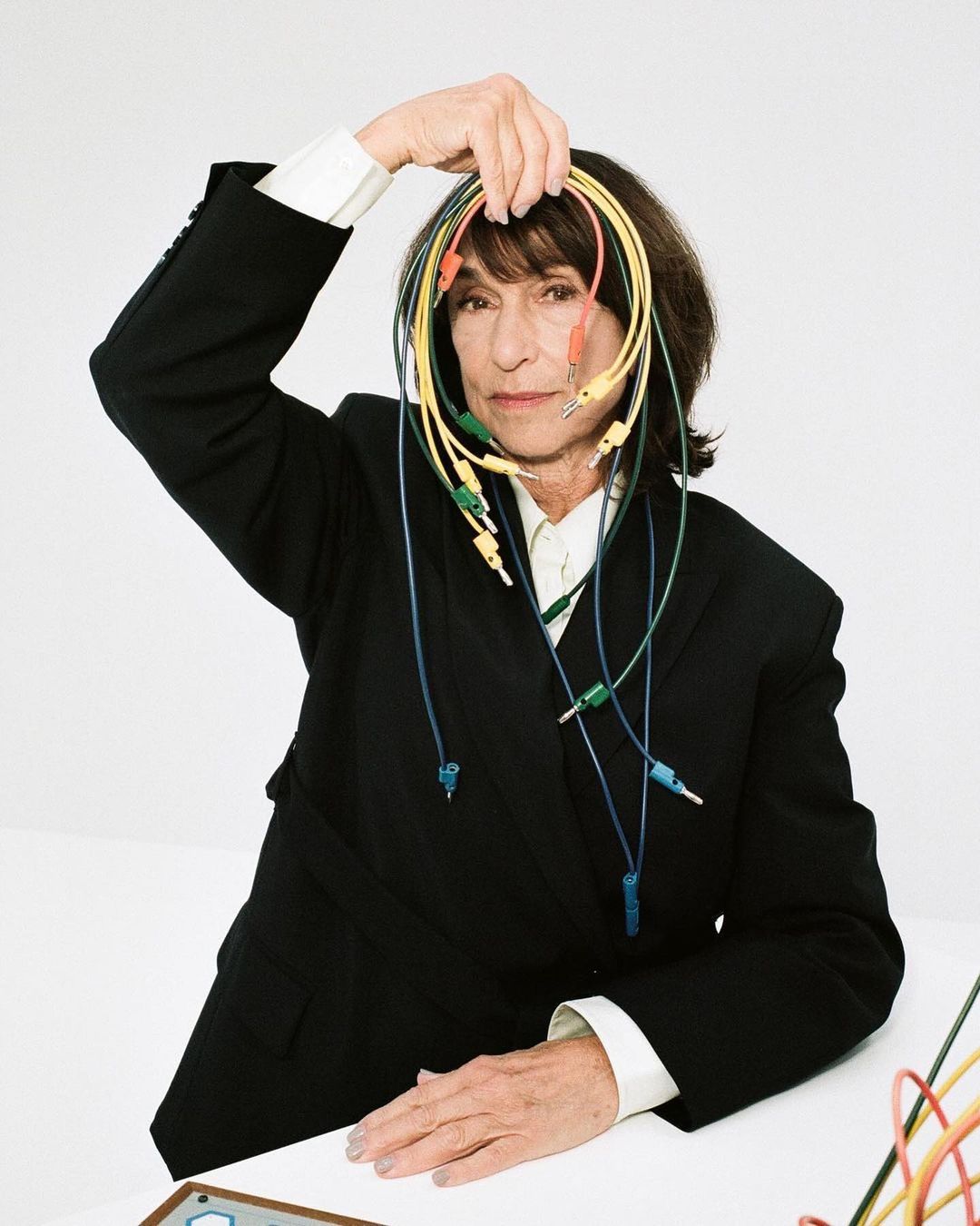Adult woman in black jacket holding an assortment of colorful electrical cables, indoors against a white background, looking at camera.