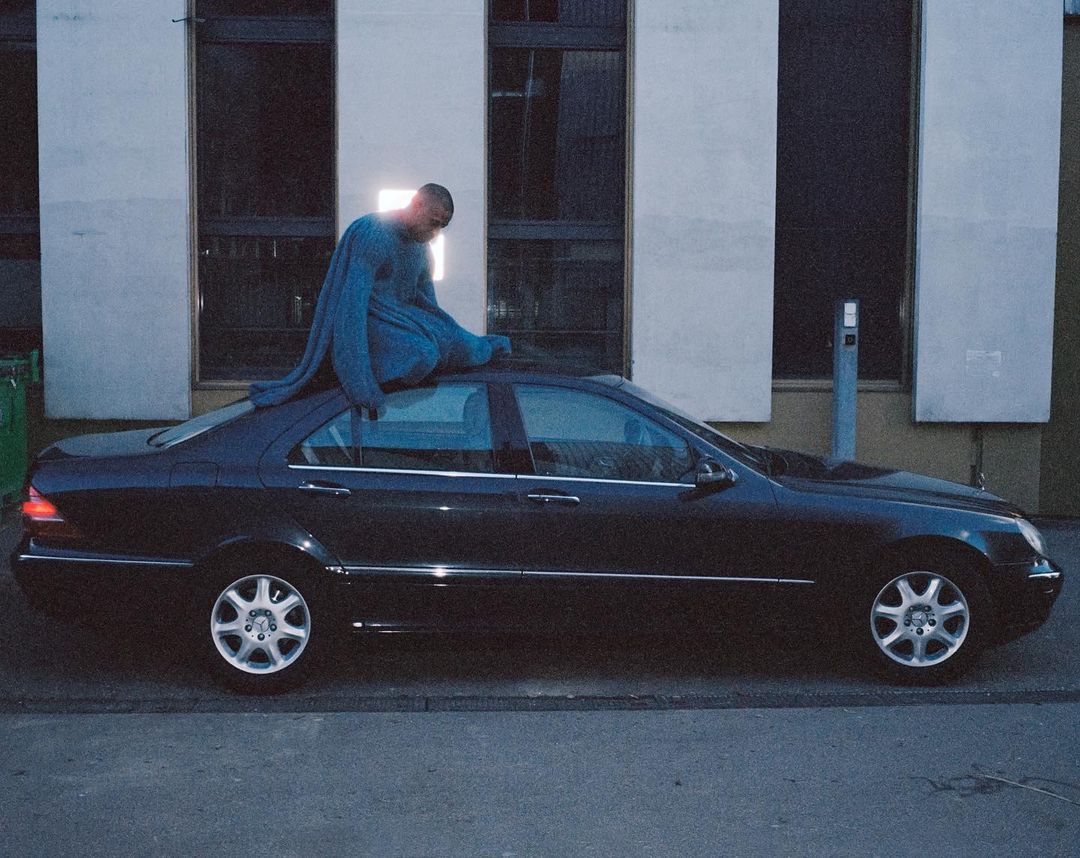 Young adult sits atop a black sedan car parked on a city street, wearing a blue sweater, next to a building with dark windows.
