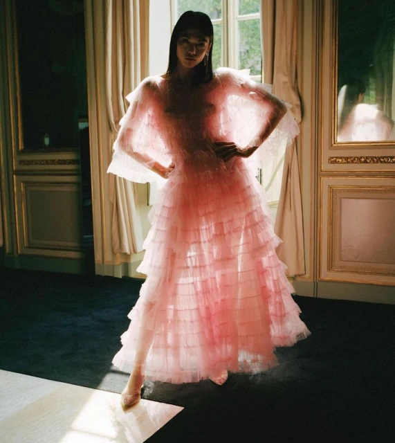 Woman in a tiered pink tulle dress posing indoors near a window with natural light creating shadows and highlights on the floor and walls.