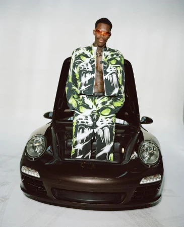 Man in graphic jacket and pants sitting on a black sportscar in a studio with white backdrop. Car hood is open showing engine.