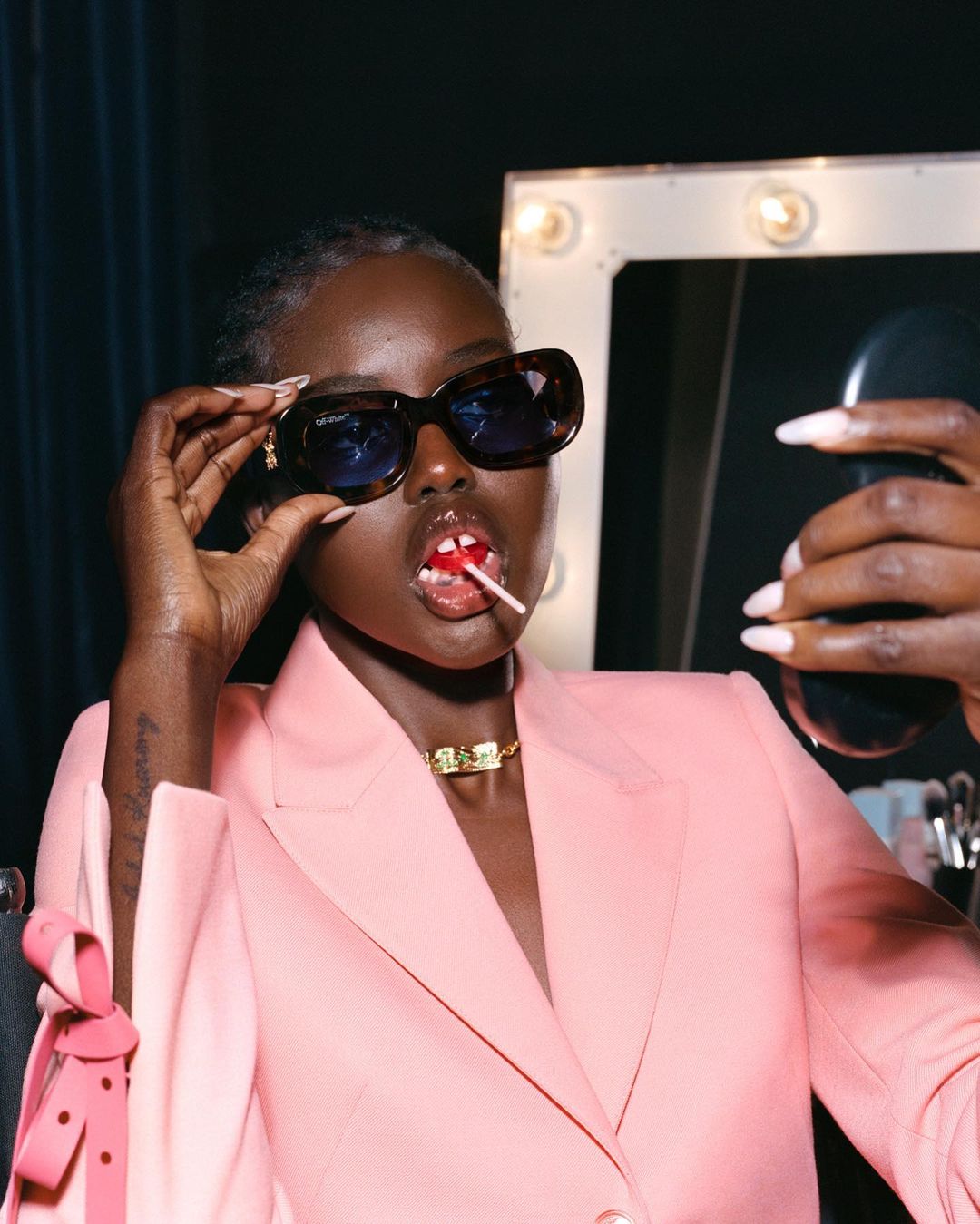 Dark-skinned woman in pink blazer and sunglasses poses with a lollipop and mirror. Makeup brushes and light-bulb mirror visible.