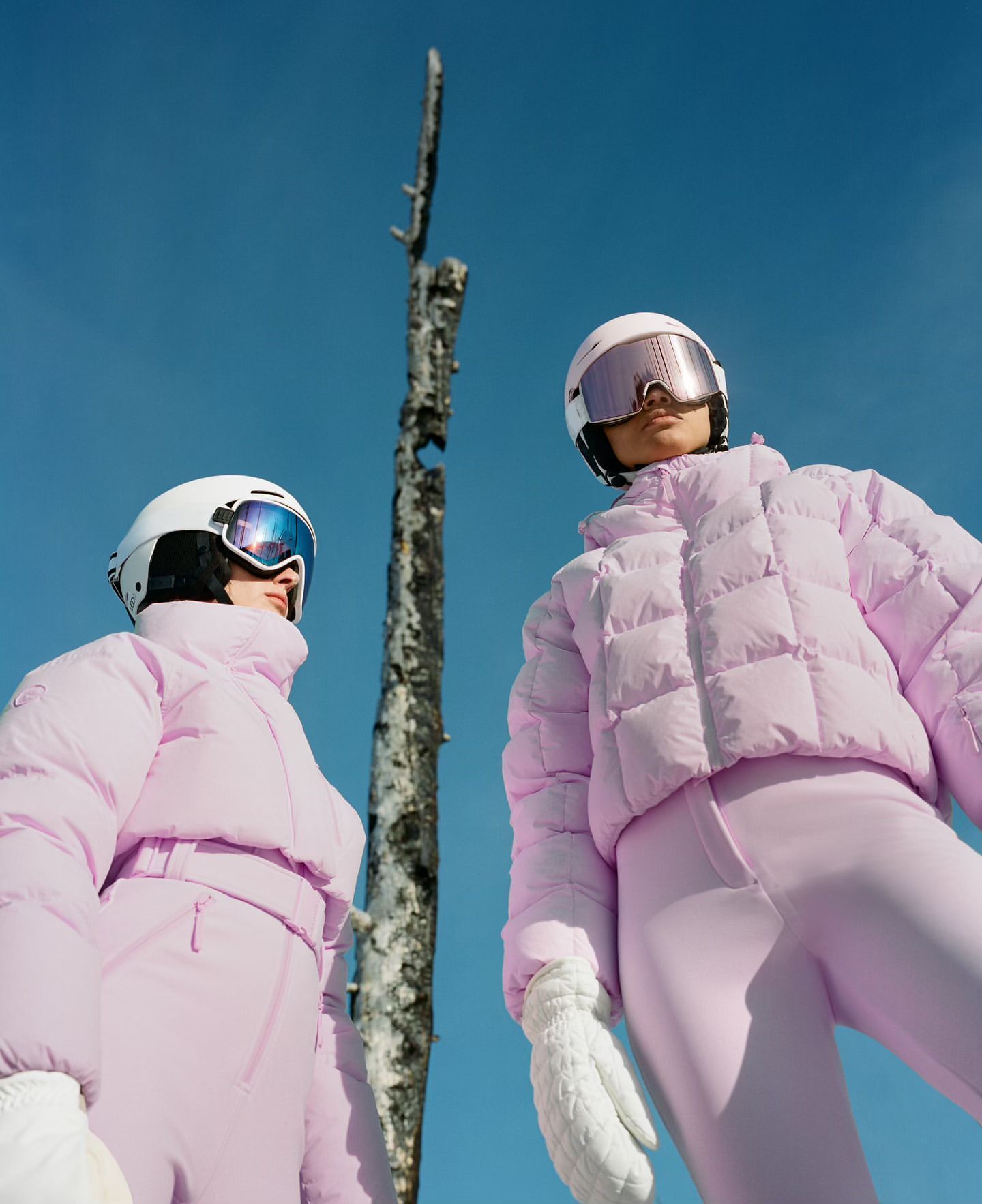 Two women in matching lavender ski suits, helmets, and goggles pose outdoors against a clear blue sky and the backdrop of a barren tree.