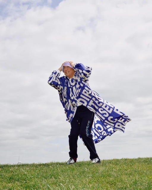 Person in patterned coat standing on a grassy hill with a cloudy sky backdrop, looking towards the horizon with hands near their head.