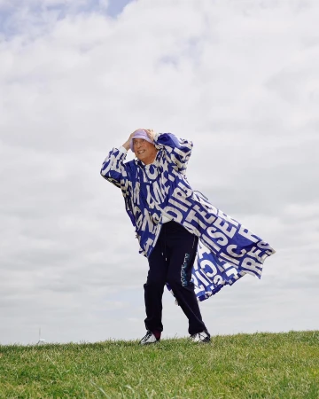 Person in patterned coat standing on a grassy hill with a cloudy sky backdrop, looking towards the horizon with hands near their head.