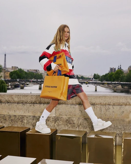 A woman walks across gold cubes in Paris, holding shopping bags, with the Eiffel Tower in the background on an overcast day.