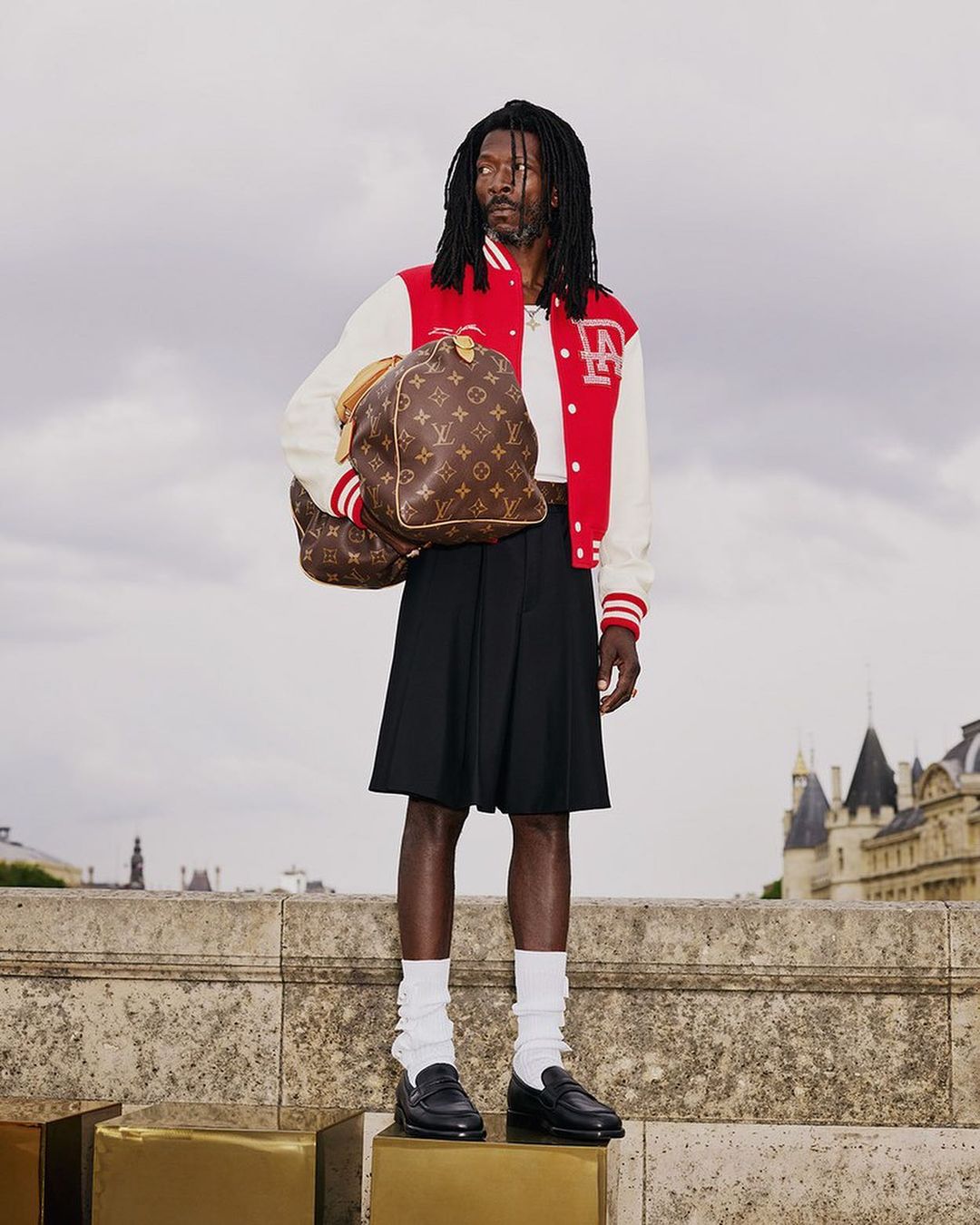 A Black male model stands on a concrete structure with a monogram travel bag, wearing a red and white jacket and black skirt in daytime.