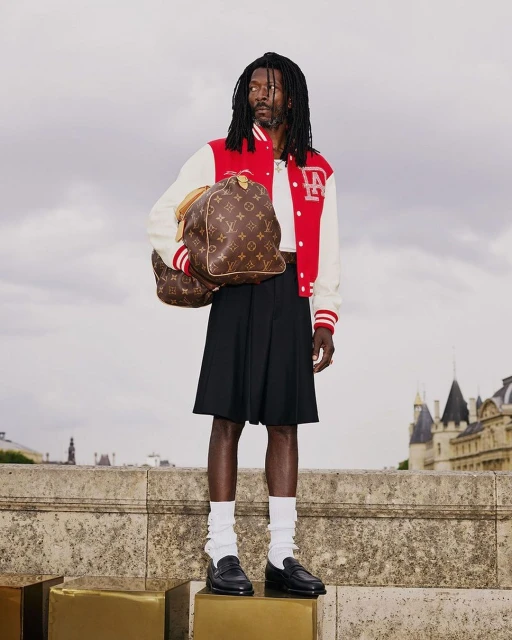 A Black male model stands on a concrete structure with a monogram travel bag, wearing a red and white jacket and black skirt in daytime.