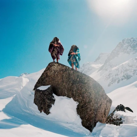 Two figures in patterned robes stand atop a rock in a snow-covered mountain landscape under a bright blue sky, creating a surreal scene.