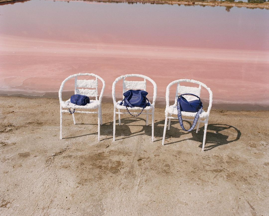 Three white chairs on a sandy beach hold blue handbags, set against a pink lake and clear sky in an outdoor fashion setting.