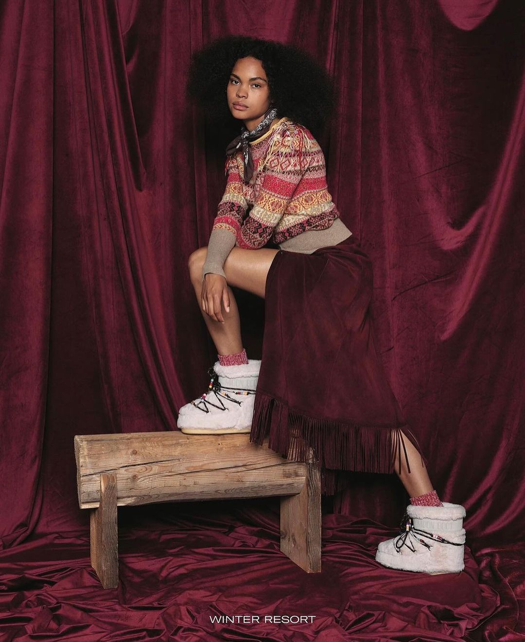 A woman poses on a wooden bench in a sweater and skirt with winter boots in front of a dark red draped fabric backdrop.