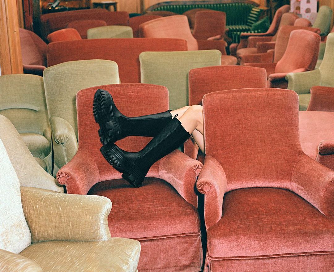 Legs of a person wearing black boots resting on a terracotta velvet armchair surrounded by many other vintage armchairs