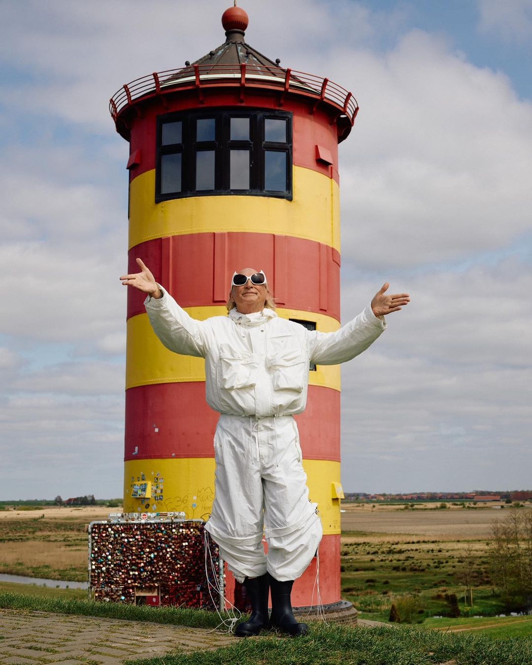 Adult male in white jumpsuit stands with arms outstretched in front of a red and yellow striped lighthouse on a grassy hill.