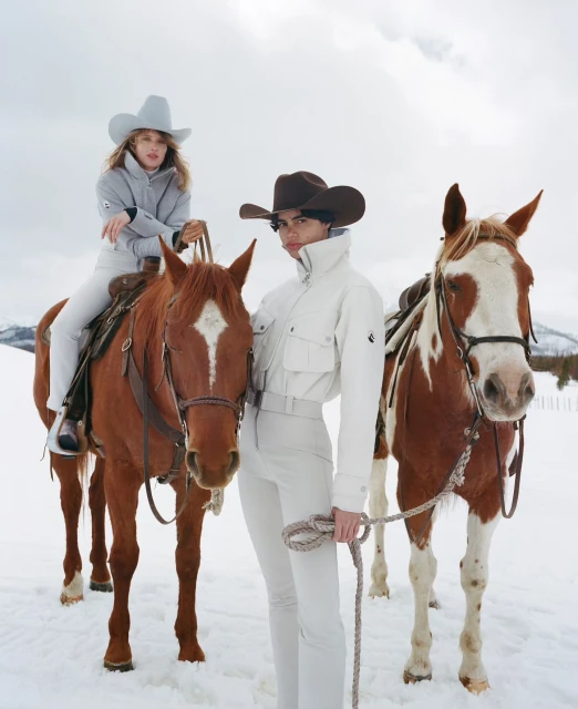 Two women in cowboy hats posing with horses in a snowy, mountainous outdoor setting, wearing winter clothing, with fair skin and light hair.