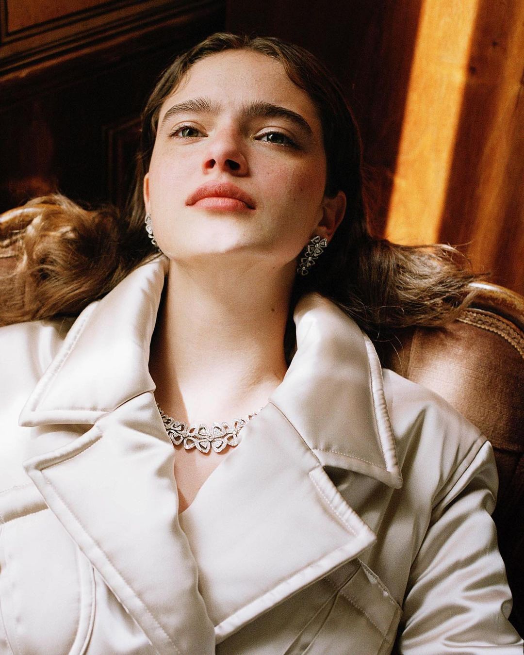 Portrait of a young woman in a white coat with elegant jewelry, seated against a dark brown background, illuminated by soft lighting.
