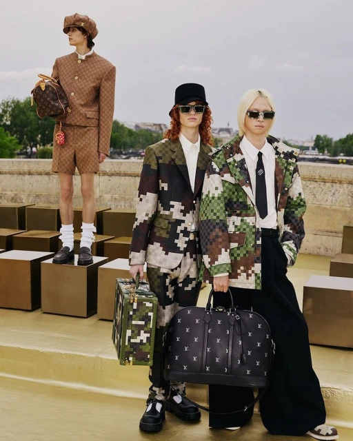 Three fashion models pose outdoors in Louis Vuitton pixelated camouflage and monogram ensembles, accessorized with handbags and luggage.