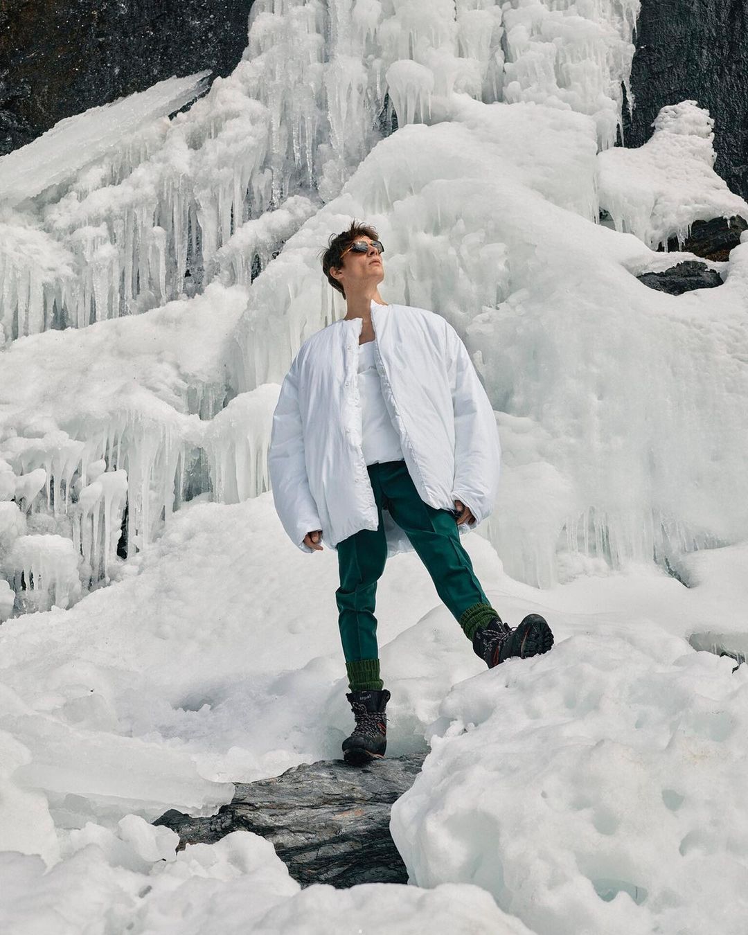 Full shot of a person in a white coat, sunglasses, and dark pants standing in snow in front of a partially frozen waterfall.