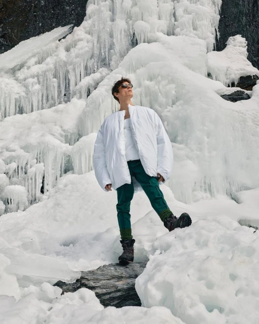 Full shot of a person in a white coat, sunglasses, and dark pants standing in snow in front of a partially frozen waterfall.
