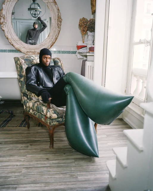 An adult in a black jacket and beanie sits in a patterned armchair with large green inflated boots in a light-filled indoor setting.